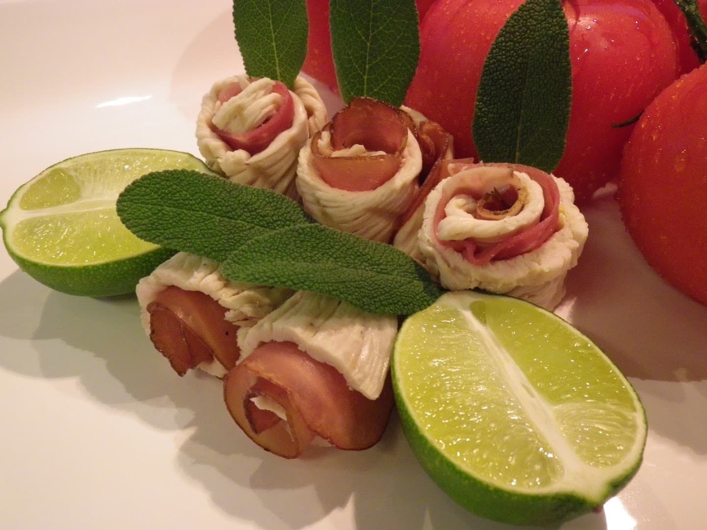 Aufgerollte Röllchen mit Salbeiblättern, halbierten Limetten und Tomaten auf weißem Teller