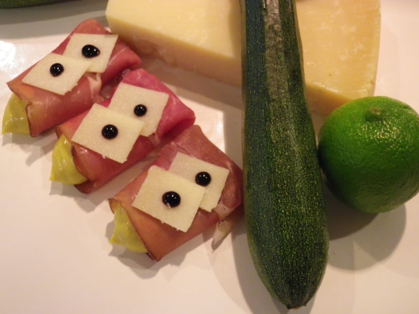 Schinkenröllchen mit Käsescheiben und Balsamico-Tupfen, daneben Zucchini, Käse und Limette